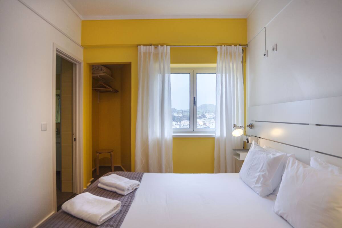 A hotel room with a bed, a window with curtains, a bedside table with a lamp, and a wardrobe.