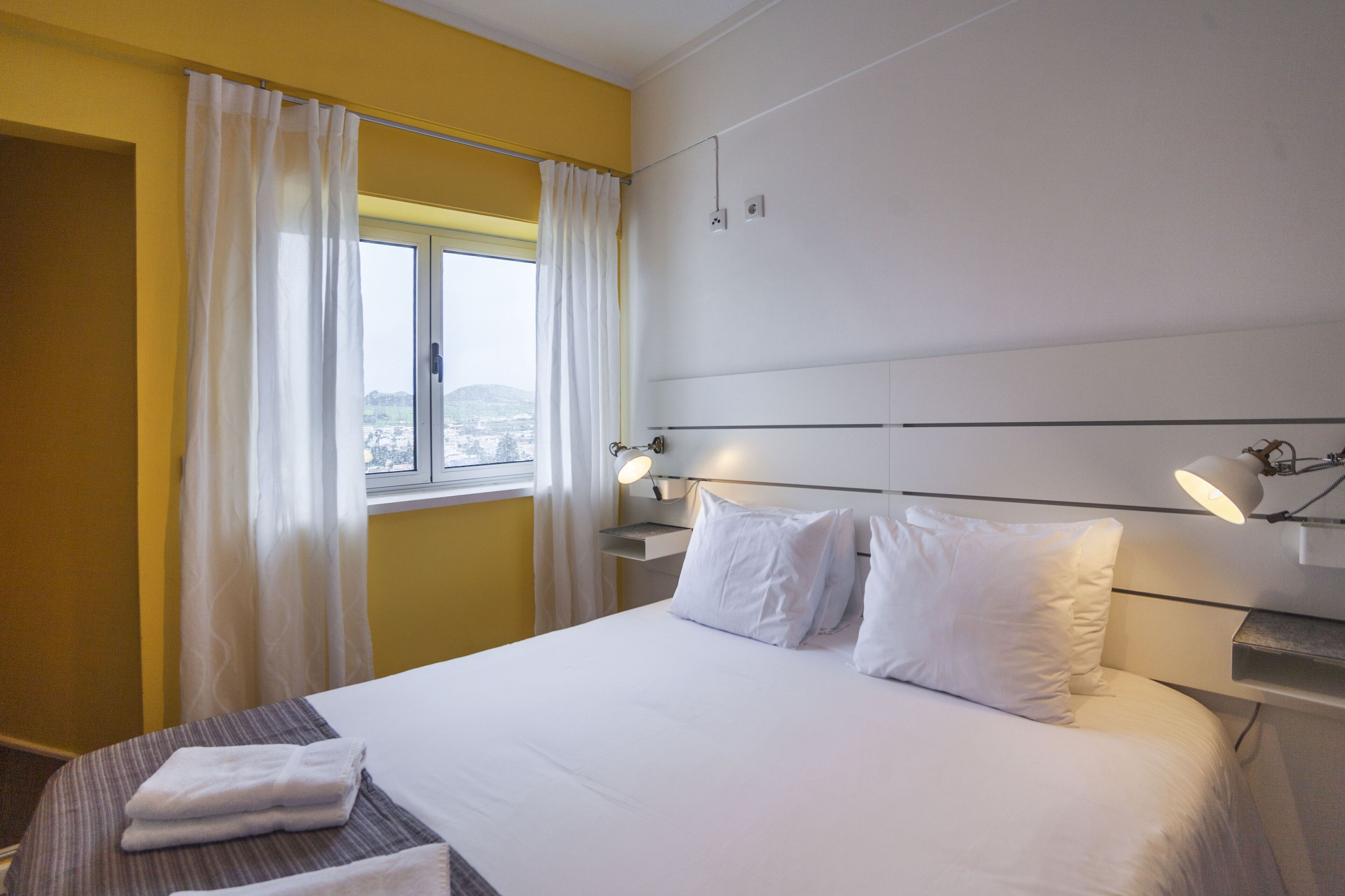 Une chambre d’hôtel avec un lit, deux lampes de chevet, une fenêtre avec des rideaux et une vue sur la ville.