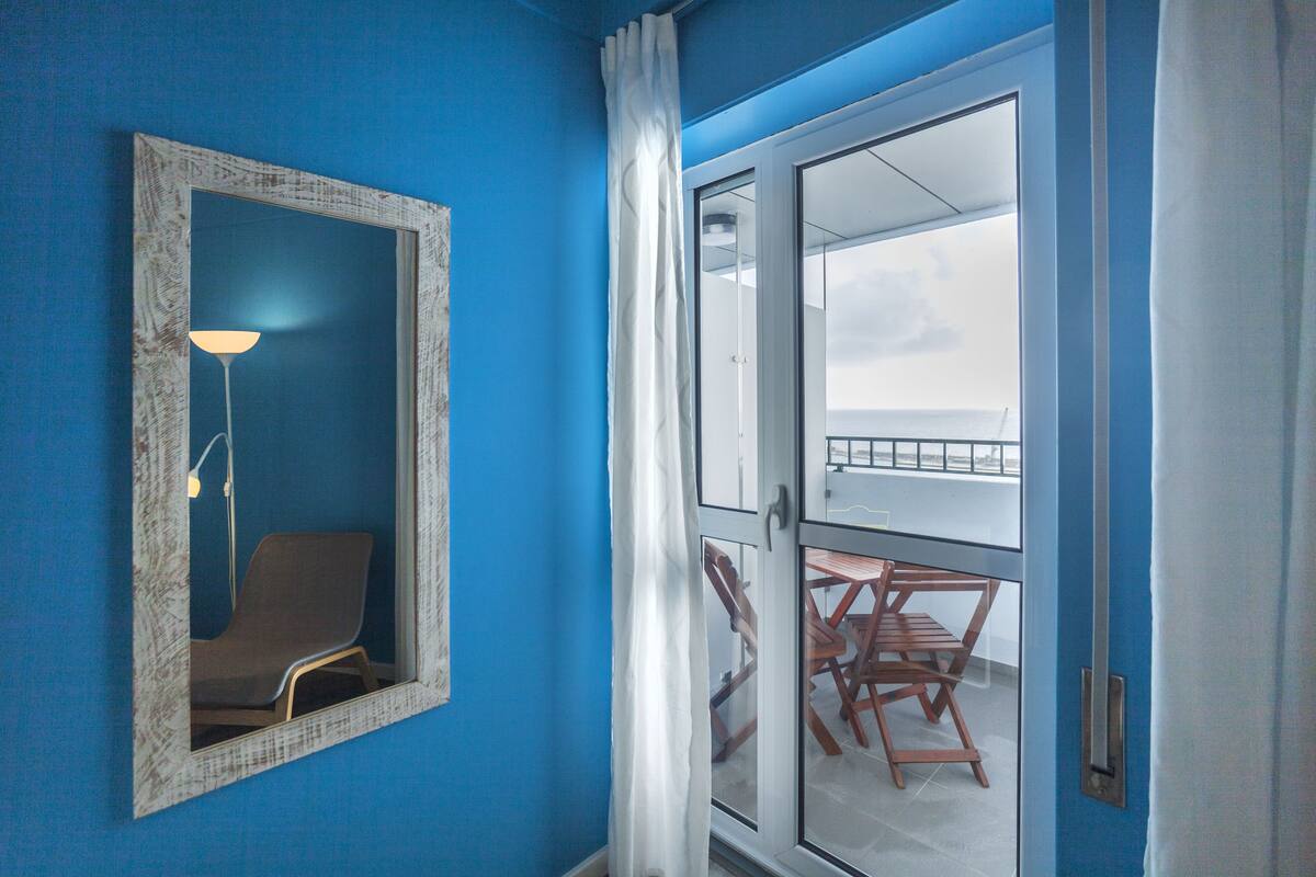 A room with a blue wall, a mirror, a chair, a table, and a view through a glass door to an outdoor balcony with a railing.