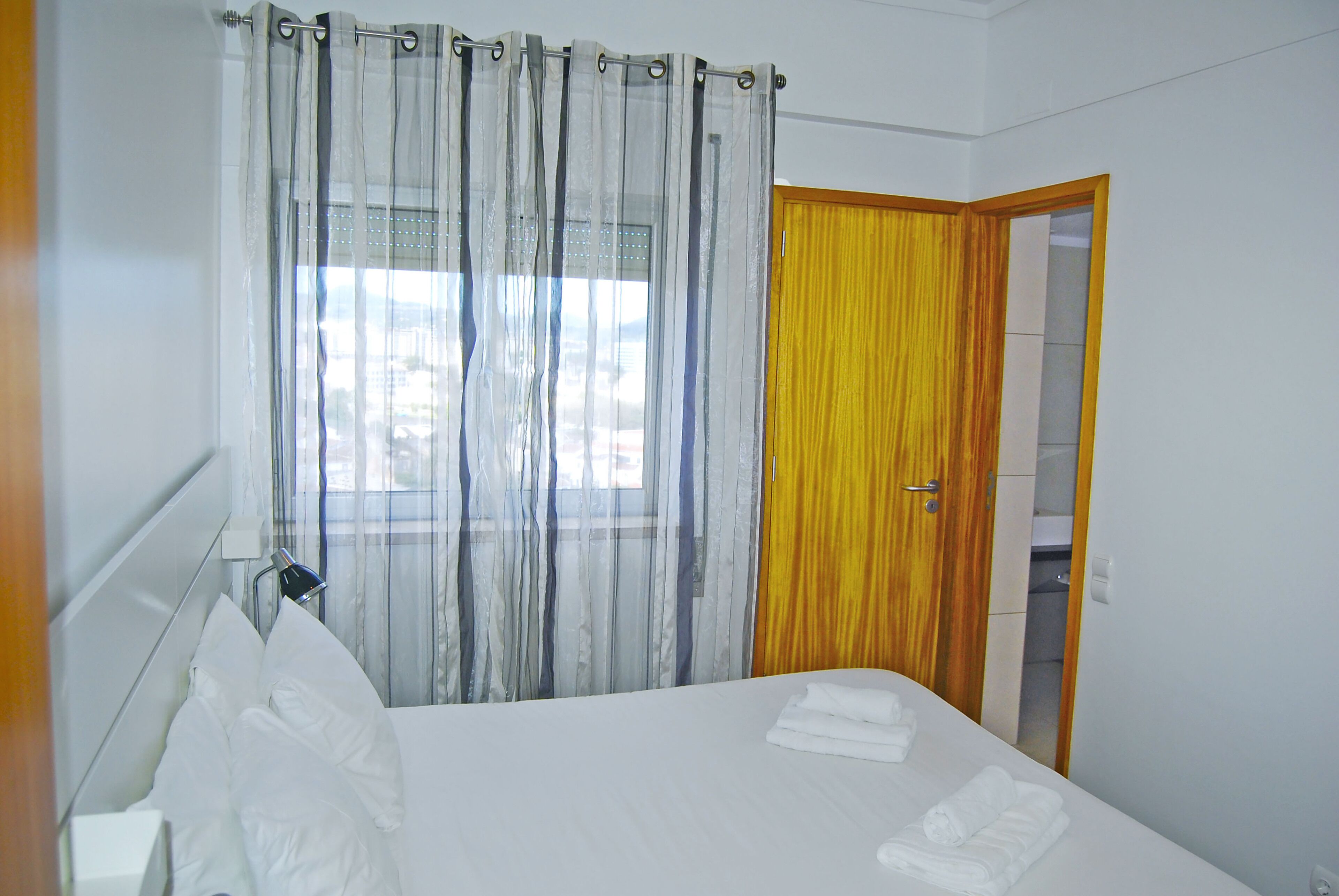 A hotel room with a bed, white curtains, a wooden door, and a window with a view.