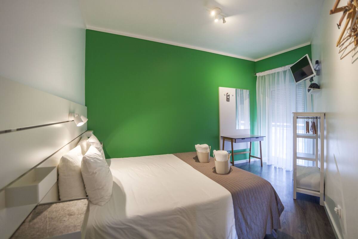 A hotel room with a green wall, a bed with white bedding, a desk with a computer, and a window with curtains.