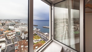 A high-rise room with a large window overlooking a coastal cityscape, featuring a view of the sea and urban buildings. - Atlantic Home Azores (Ponta Delgada)