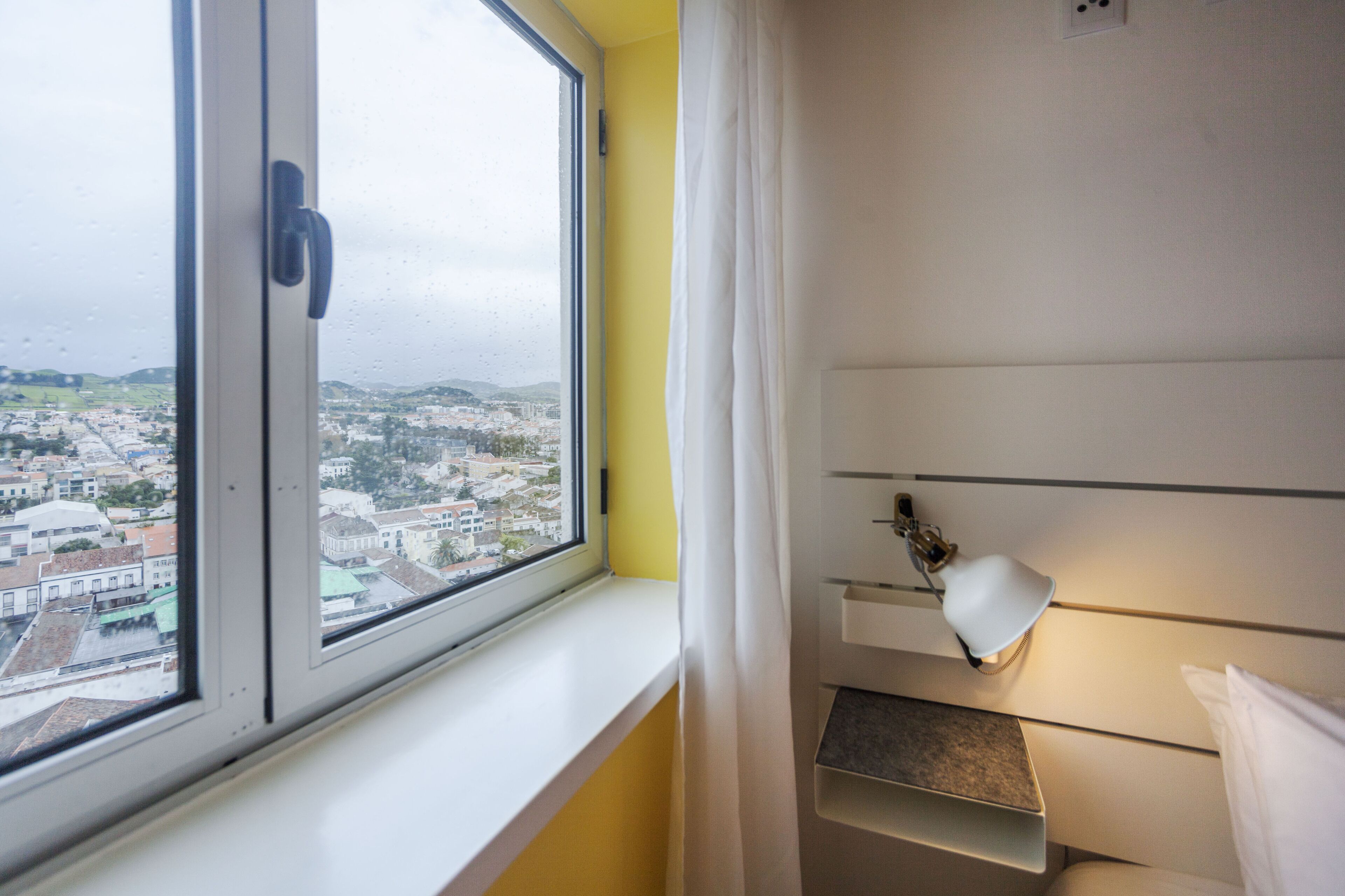 Een hotelkamer met een raam dat uitkijkt op een stadsgezicht, een bedlampje en een wit gordijn.