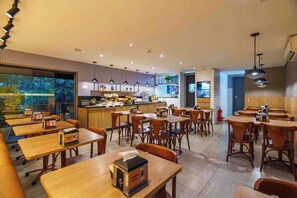 A modern restaurant with wooden tables and chairs, a kitchen area, and a view of trees through a glass wall. - Uniclass Hotel Pinheiros (São Paulo)