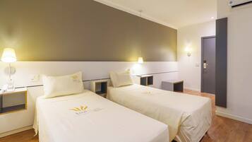 A hotel room with two beds, each with a white bedspread and matching pillows, a bedside table with a lamp, and a wall-mounted light fixture.