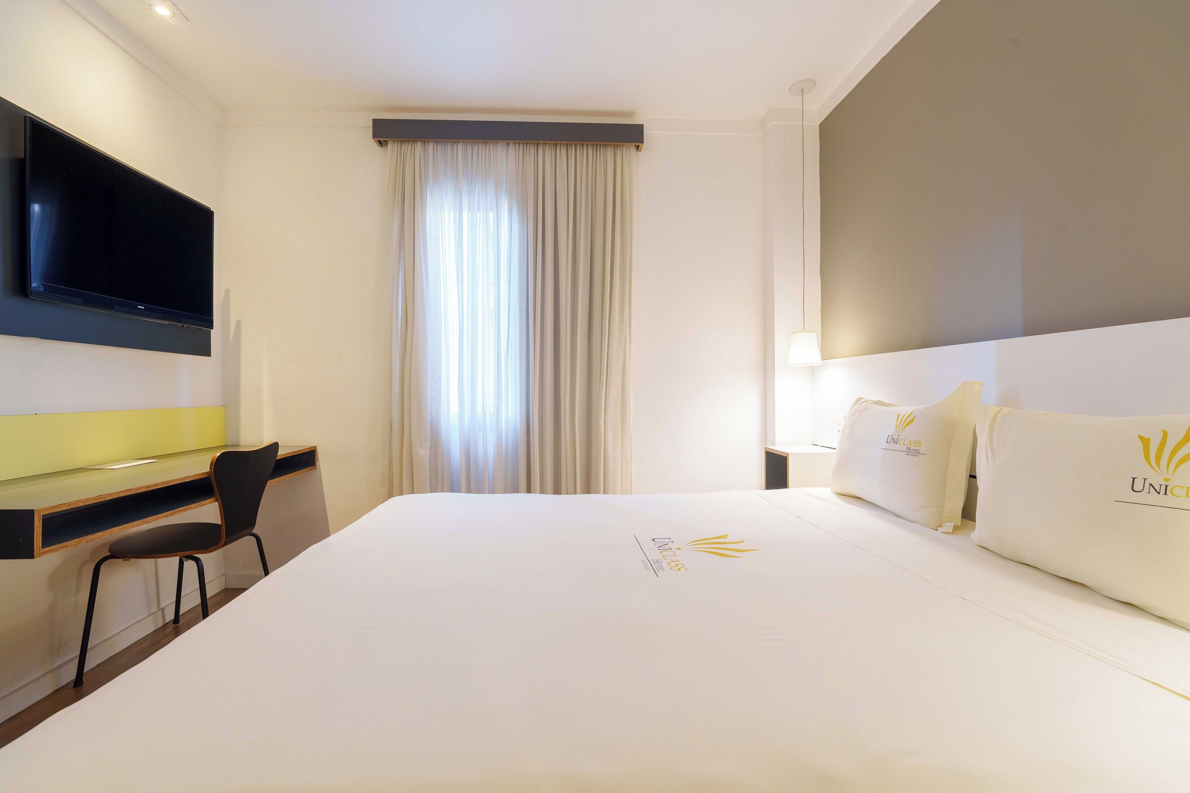 Ein Hotelzimmer mit Bett, Schreibtisch, Stuhl, einem an der Wand befestigten Fernseher und einem Fenster mit Vorhängen.