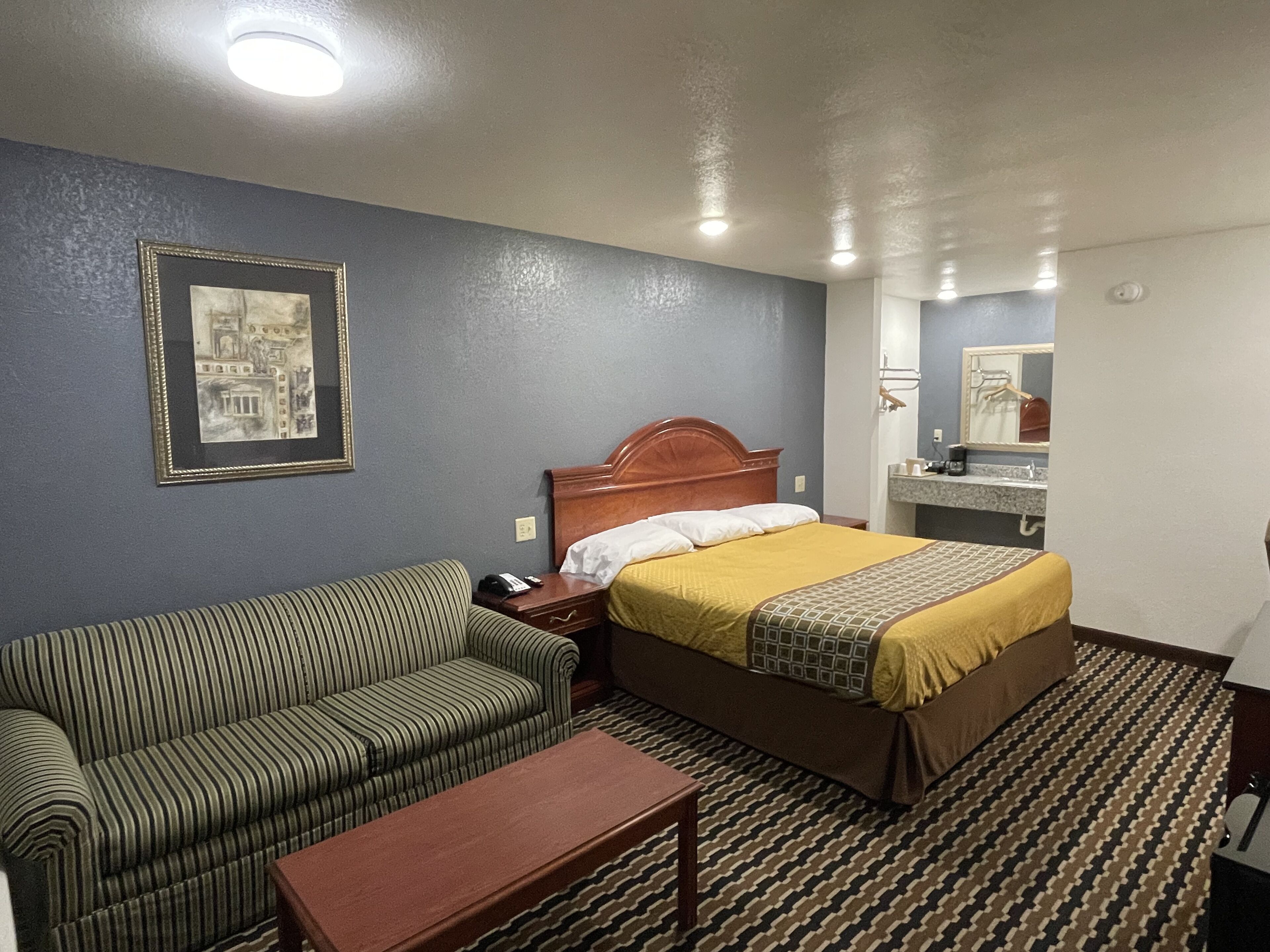 Una habitación de hotel con una cama, un sofá a rayas, una mesita de noche de madera, un cuadro en la pared y un baño visible a través de una puerta abierta.