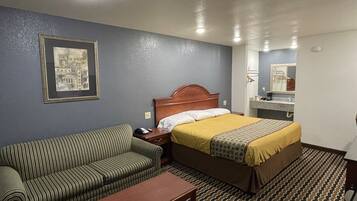 Una habitación de hotel con una cama, un sofá a rayas, una mesita de noche de madera, un cuadro en la pared y un baño visible a través de una puerta abierta.