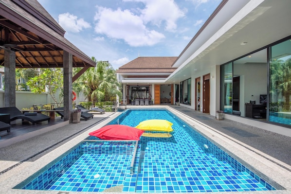 A swimming pool with lounge chairs and umbrellas, a covered area, and a building with large windows.