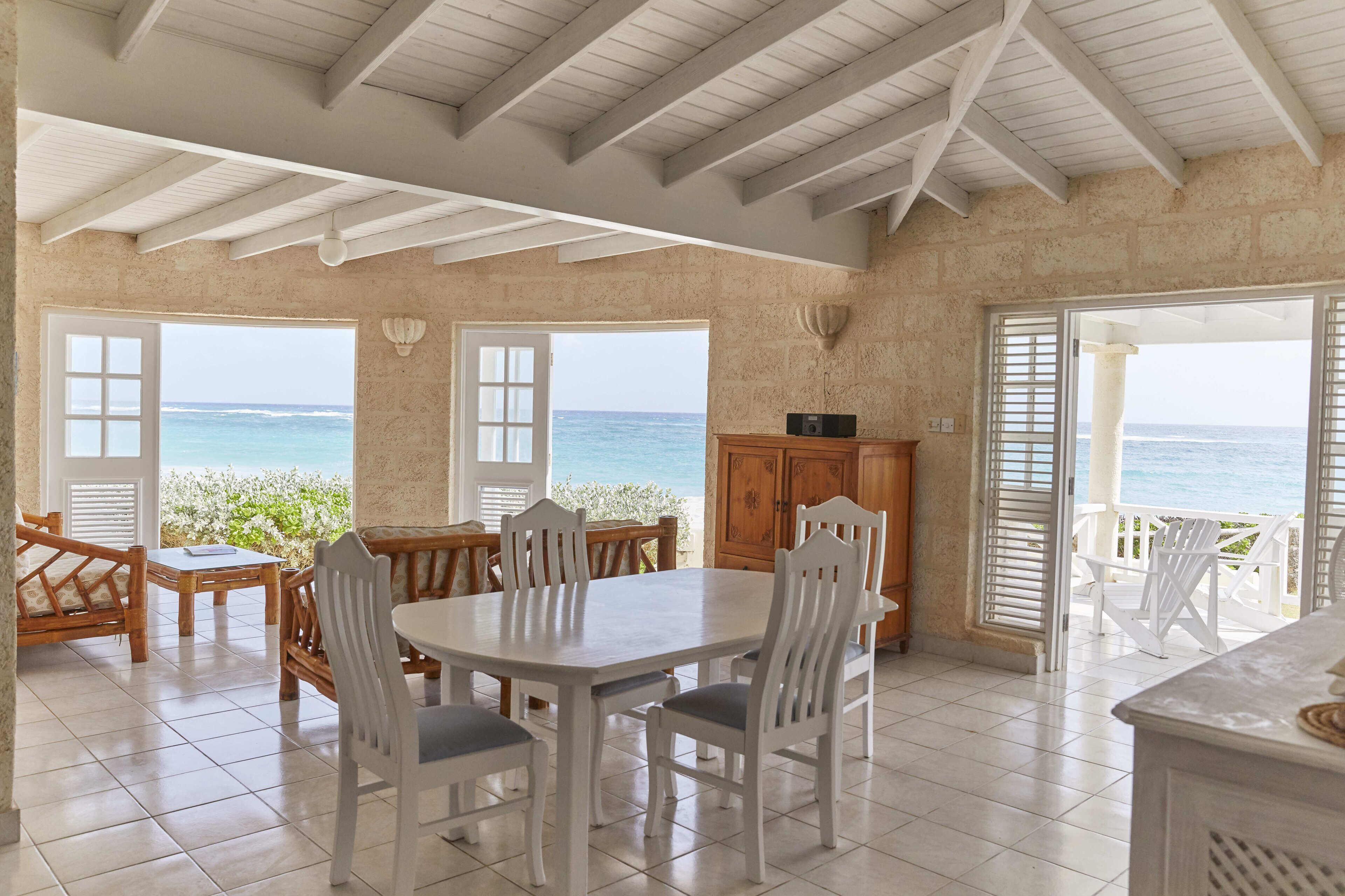 A dining area with a view of the sea, featuring a large table, chairs, and a wooden cabinet.