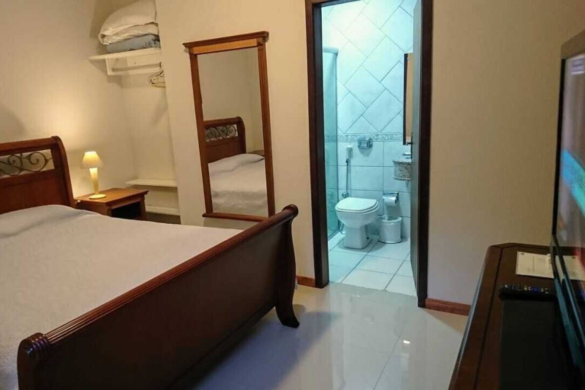 A hotel room with a bed, a bedside table with a lamp, a mirror, a television, and a bathroom visible through an open door.
