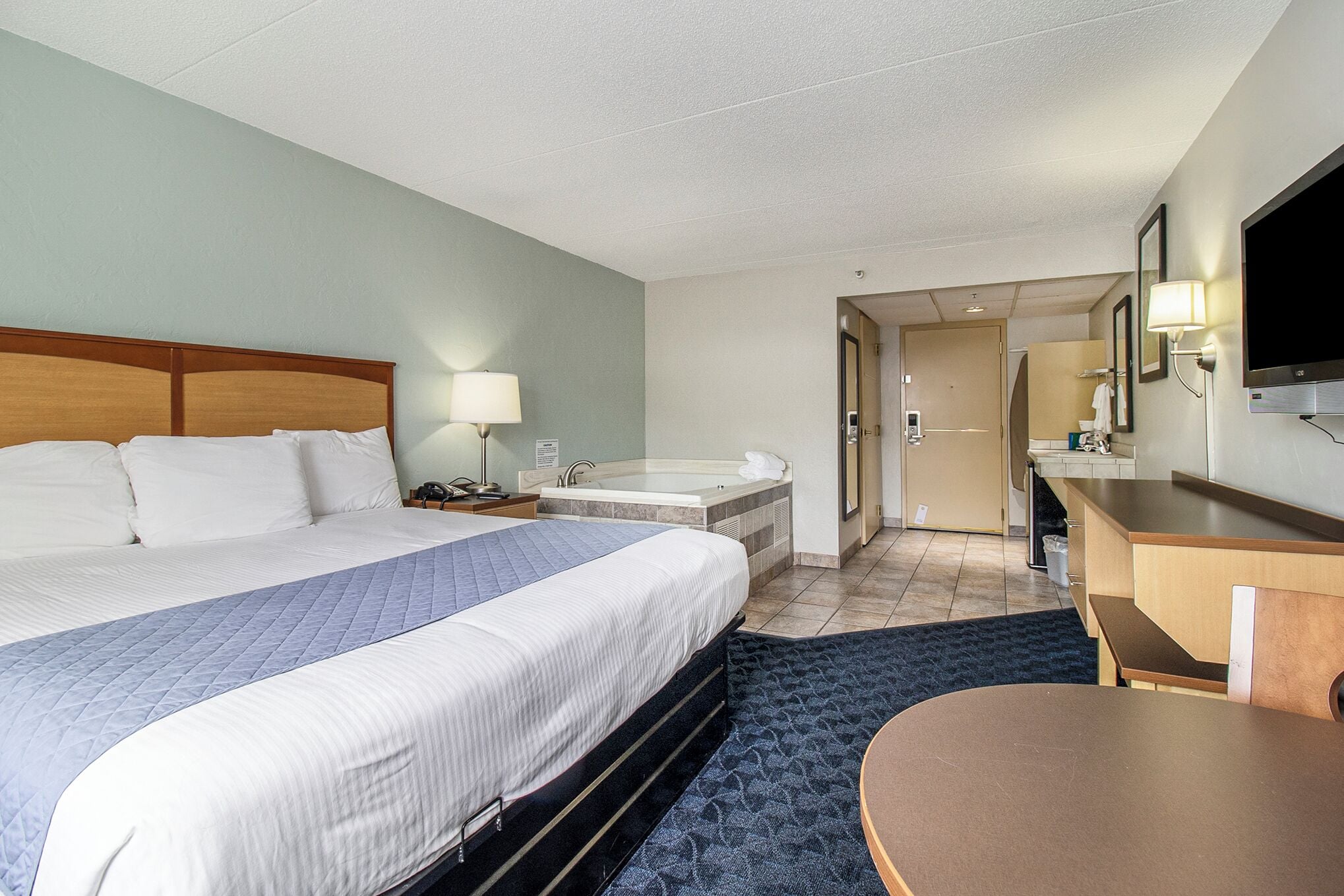 Una habitación de hotel con una cama grande, un escritorio, una silla, una lámpara de escritorio, un televisor, un baño con bañera y un espejo.