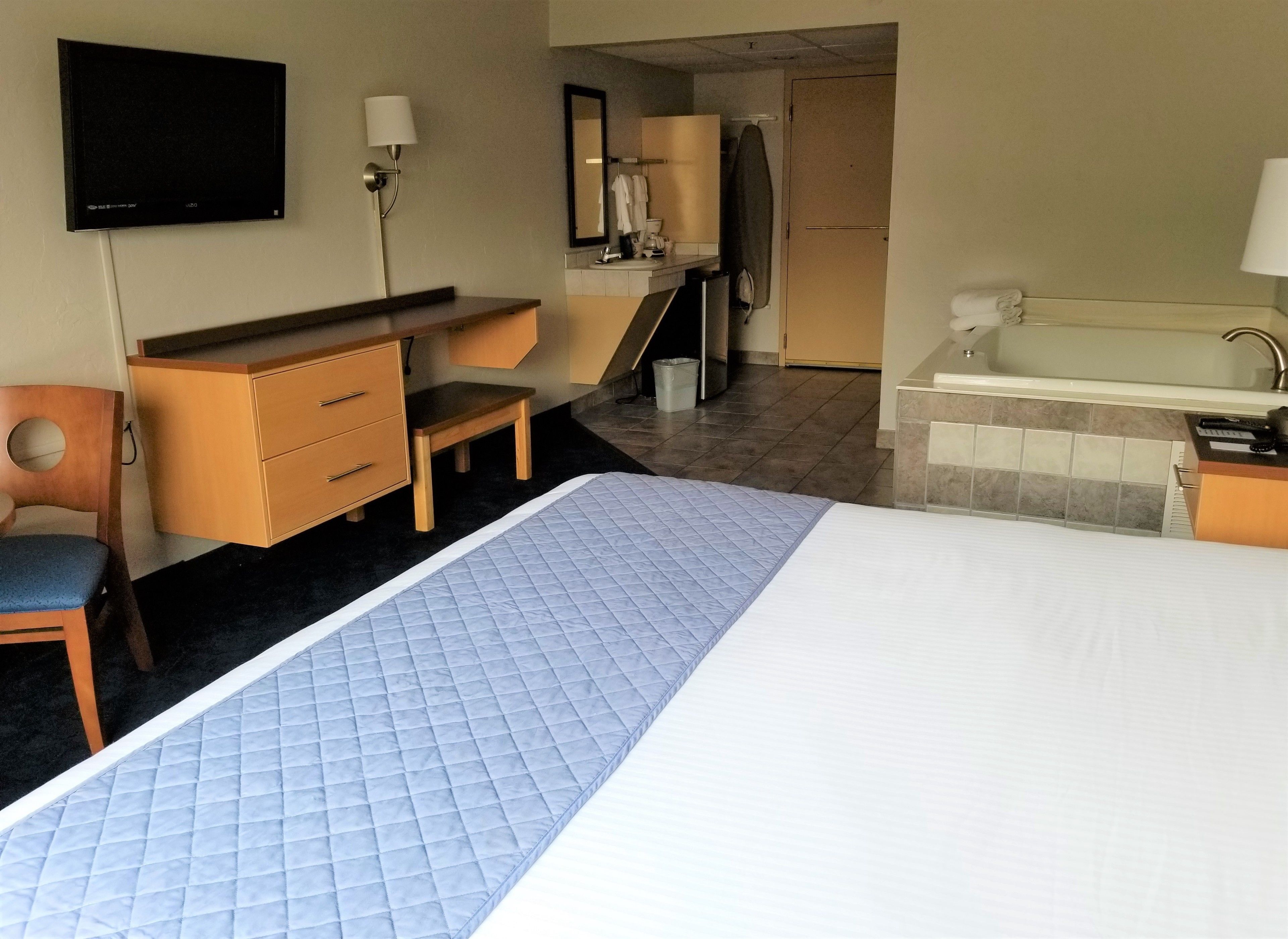 A hotel room with a bed, a desk, a chair, a small table, a mirror, a sink, and a television.