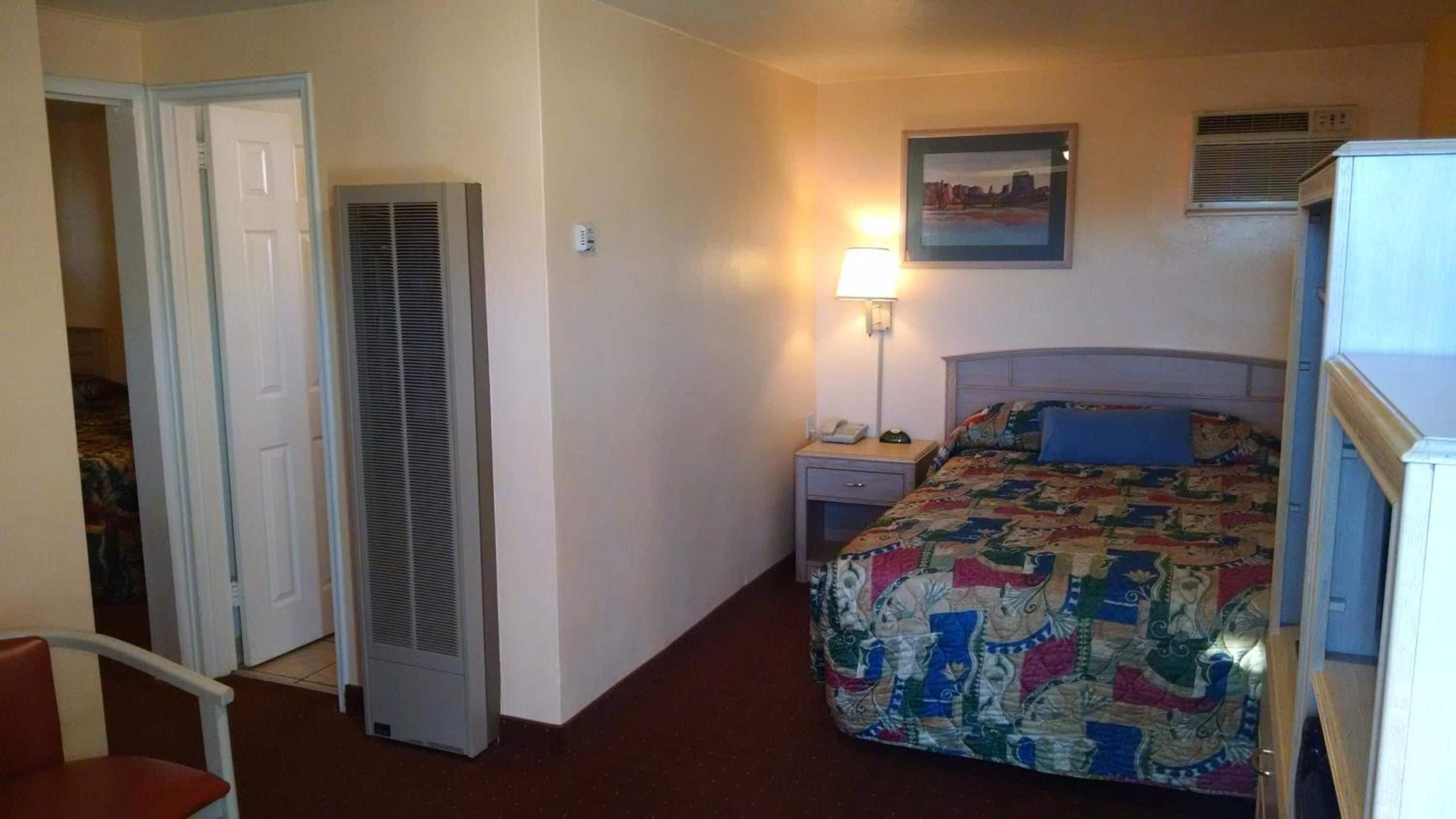 A hotel room with a bed, a bedside table with a lamp, a chair, and a door leading to another room.