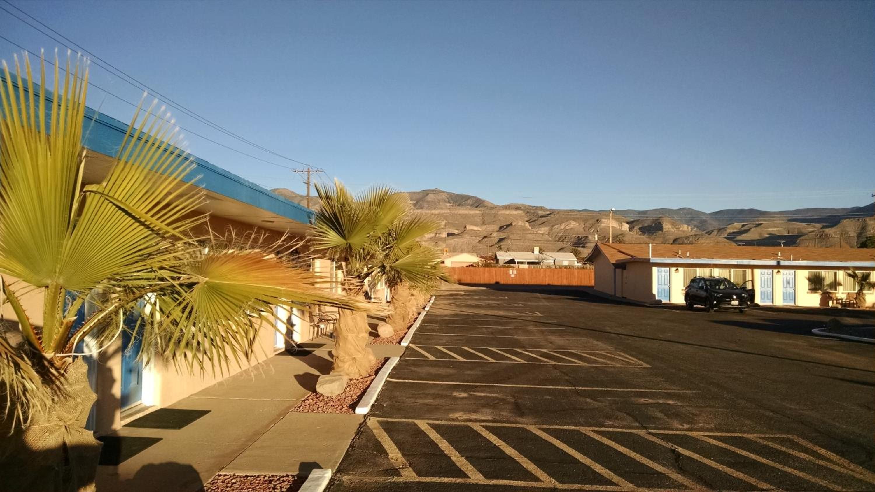 Ein Parkplatz mit einem Gebäude links, einer Palme und einem Gebirge im Hintergrund.