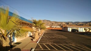 A parking lot with a building on the left, a palm tree, and a mountain range in the background. - White Sands Motel (Alamogordo)