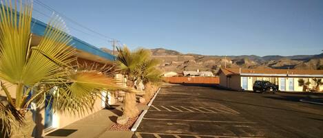 A parking lot with a building on the left, a palm tree, and a mountain range in the background.