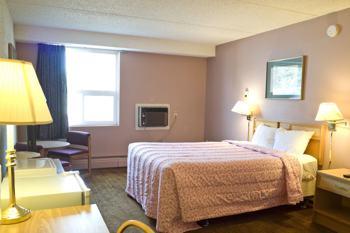 A hotel room with a bed, a desk, a chair, a table, a lamp, a window, and a picture on the wall.