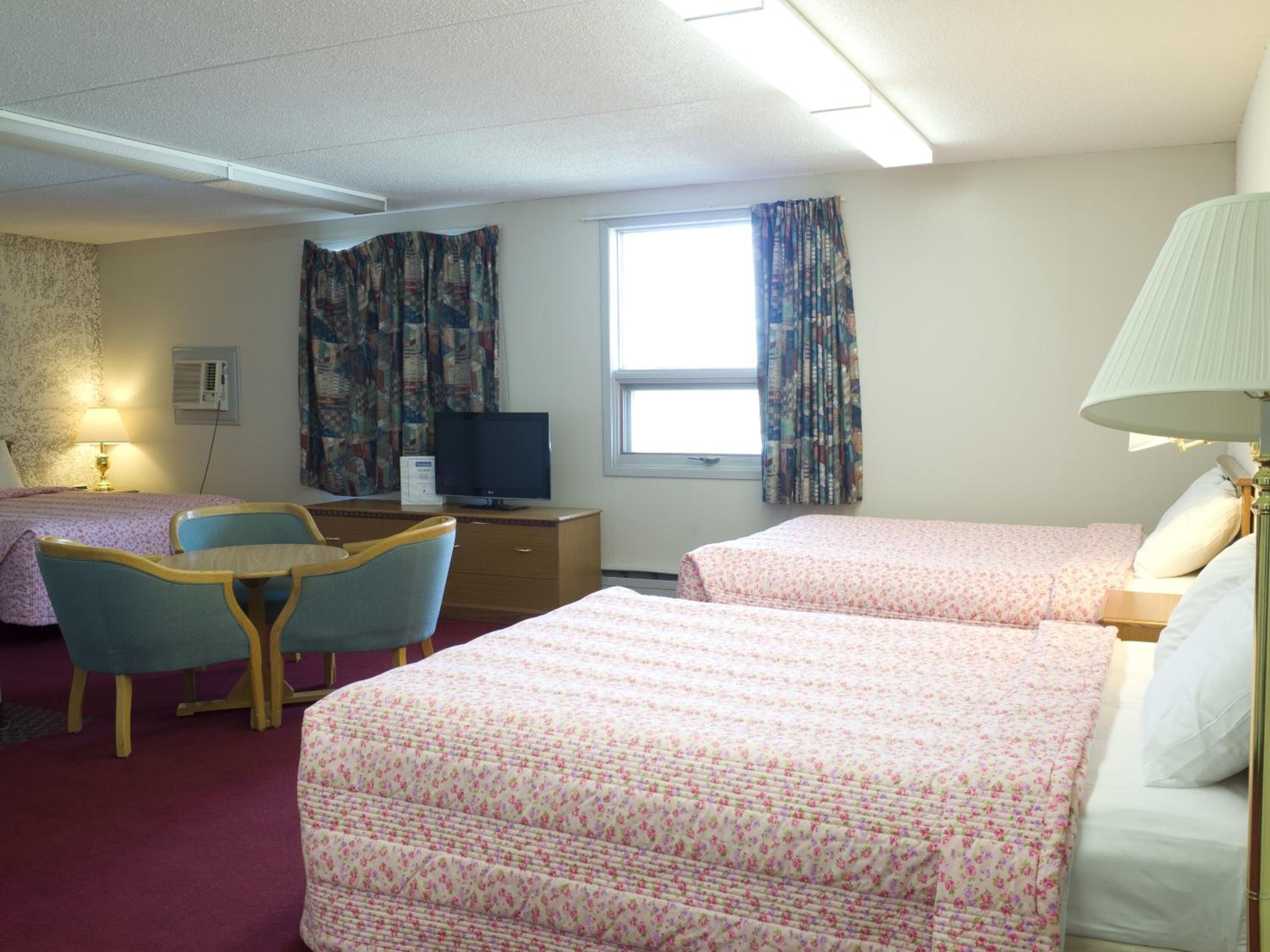 Quarto de hotel com duas camas, uma escrivaninha com computador, uma cadeira, um abajur e uma janela com cortinas.