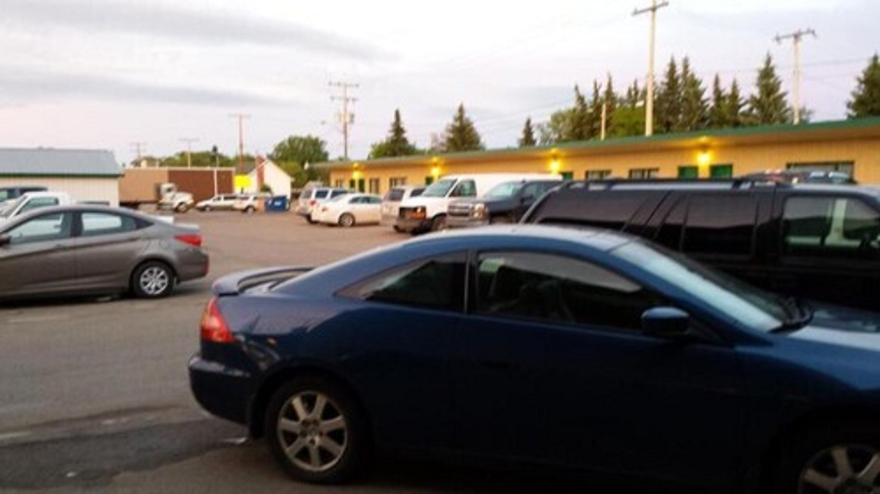 A parking lot with several cars, a building with a green roof, and a few trees in the background.