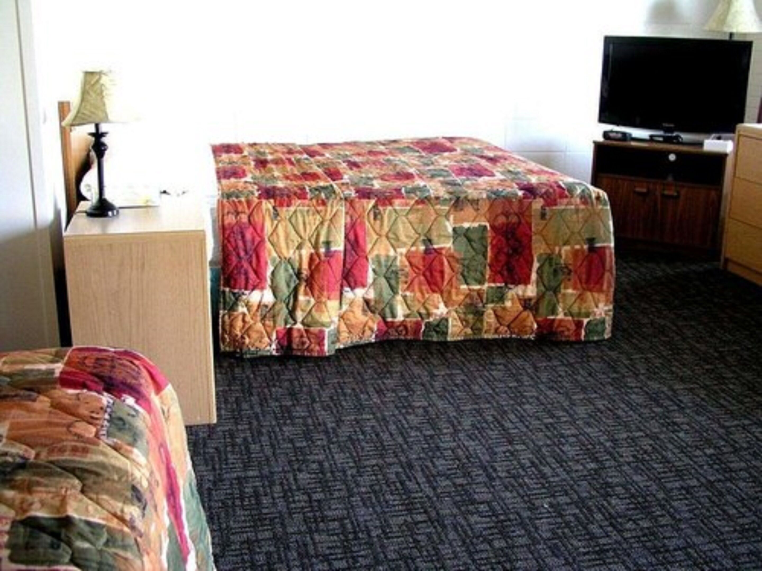 A hotel room with a bed covered in a colorful quilt, a television on a wooden stand, and a carpeted floor.