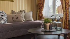 Una sala de estar acogedora con un sofá, cojines, una mesa redonda con velas y una planta, y una ventana con cortinas.