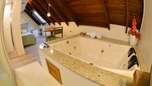 A modern bathroom with a large bathtub, wooden ceiling beams, and a small seating area with a chair and table. - Hotel Renascença (Gramado)