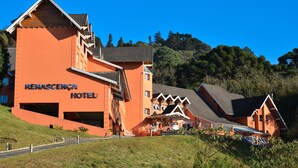 A large, orange building with the name "Renascença Hotel" prominently displayed. It features multiple levels, a sloped roof, and a parking area in front. - Hotel Renascença (Gramado)