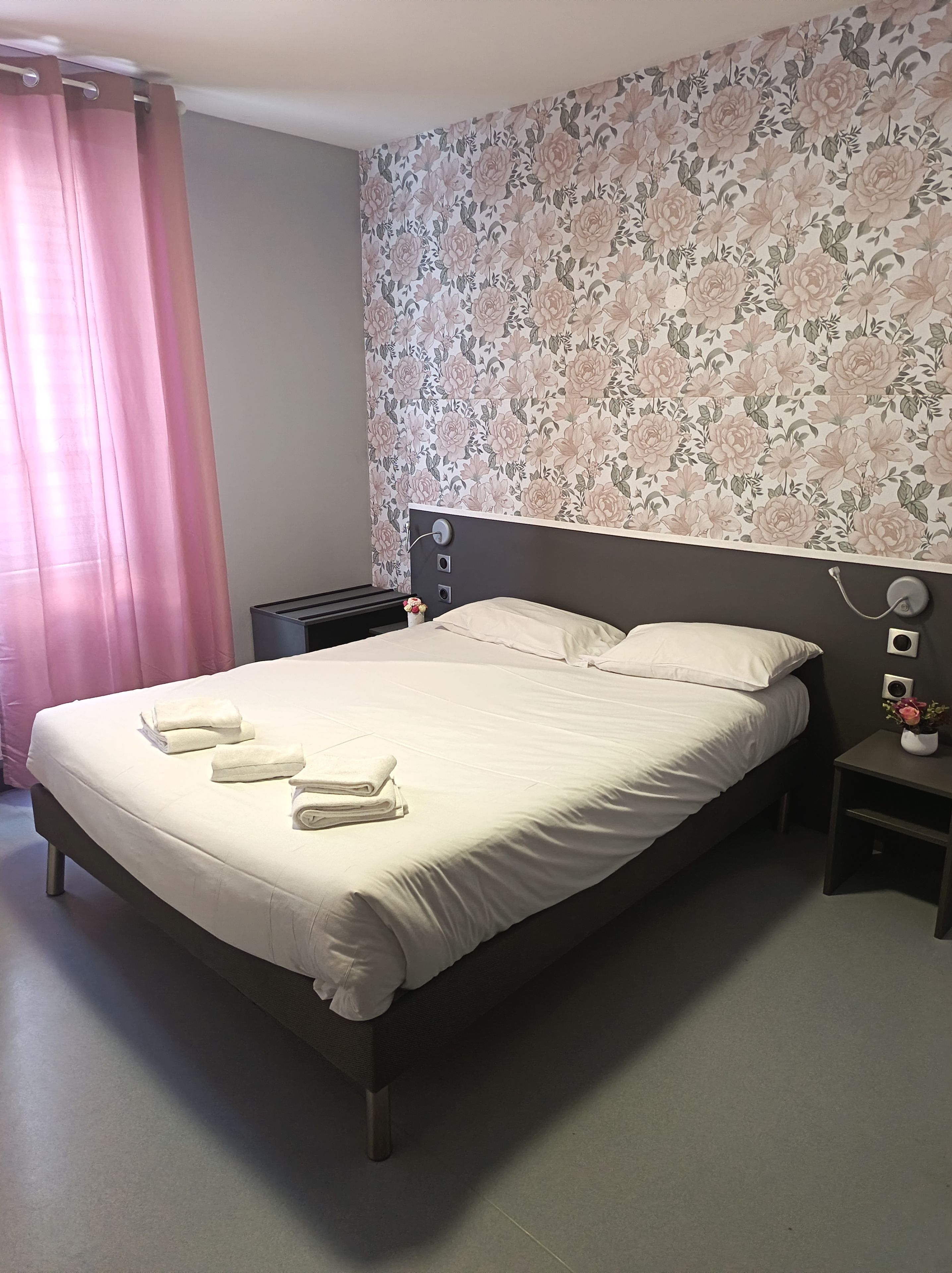 A neatly made bed with white linens and towels, a dark headboard, a nightstand with a lamp, and a floral wallpaper pattern on the wall.
