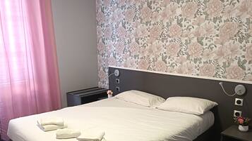 A neatly made bed with white linens and towels, a dark headboard, a nightstand with a lamp, and a floral wallpaper pattern on the wall.