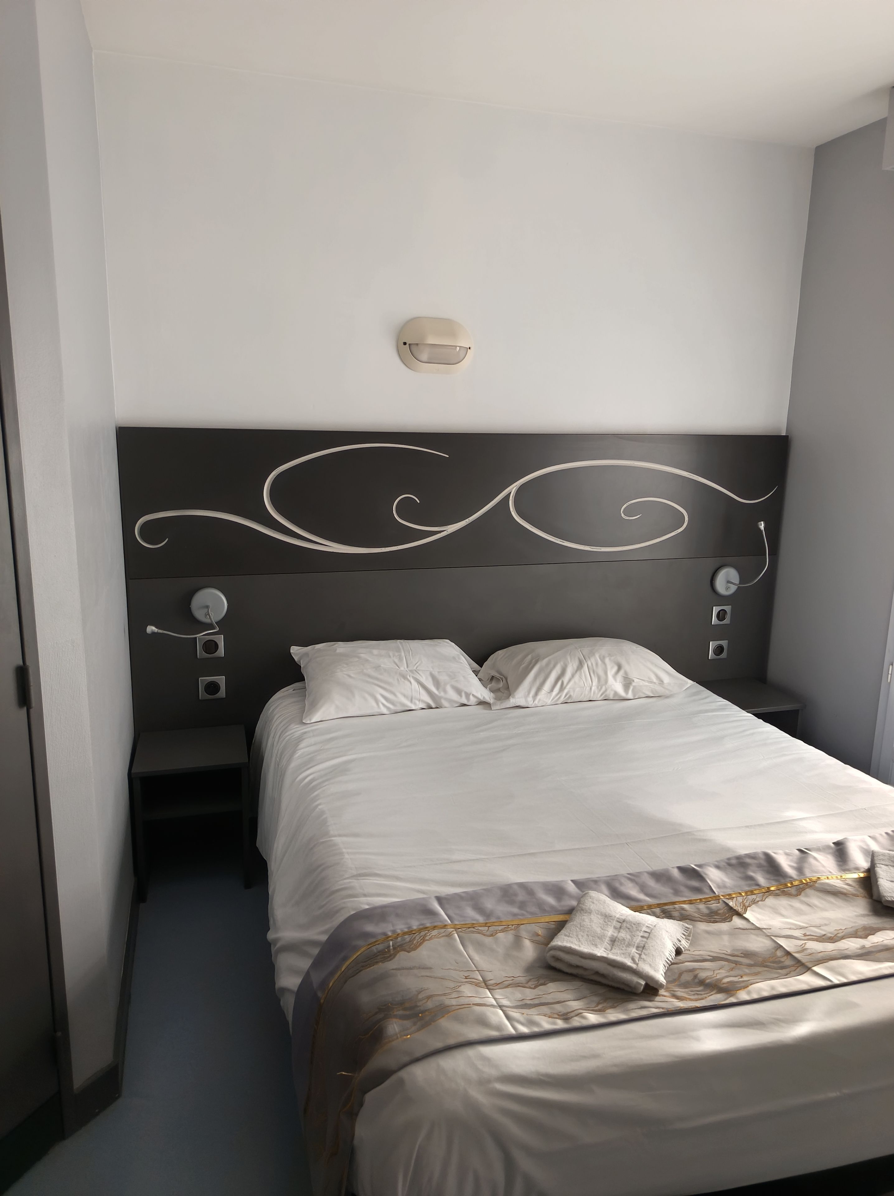 A hotel room with a bed, bedside tables, and a headboard with decorative swirls.