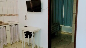 Una habitación pequeña con una televisión montada en la pared, una mesa blanca con una silla y un baño visible a través de un puerta abierta.