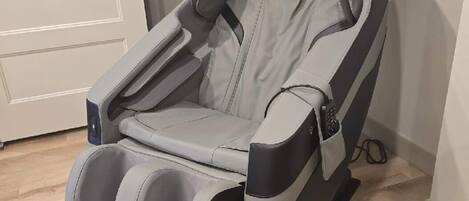 A massage chair with a footrest, positioned in a room with a wooden floor and a white door in the background.