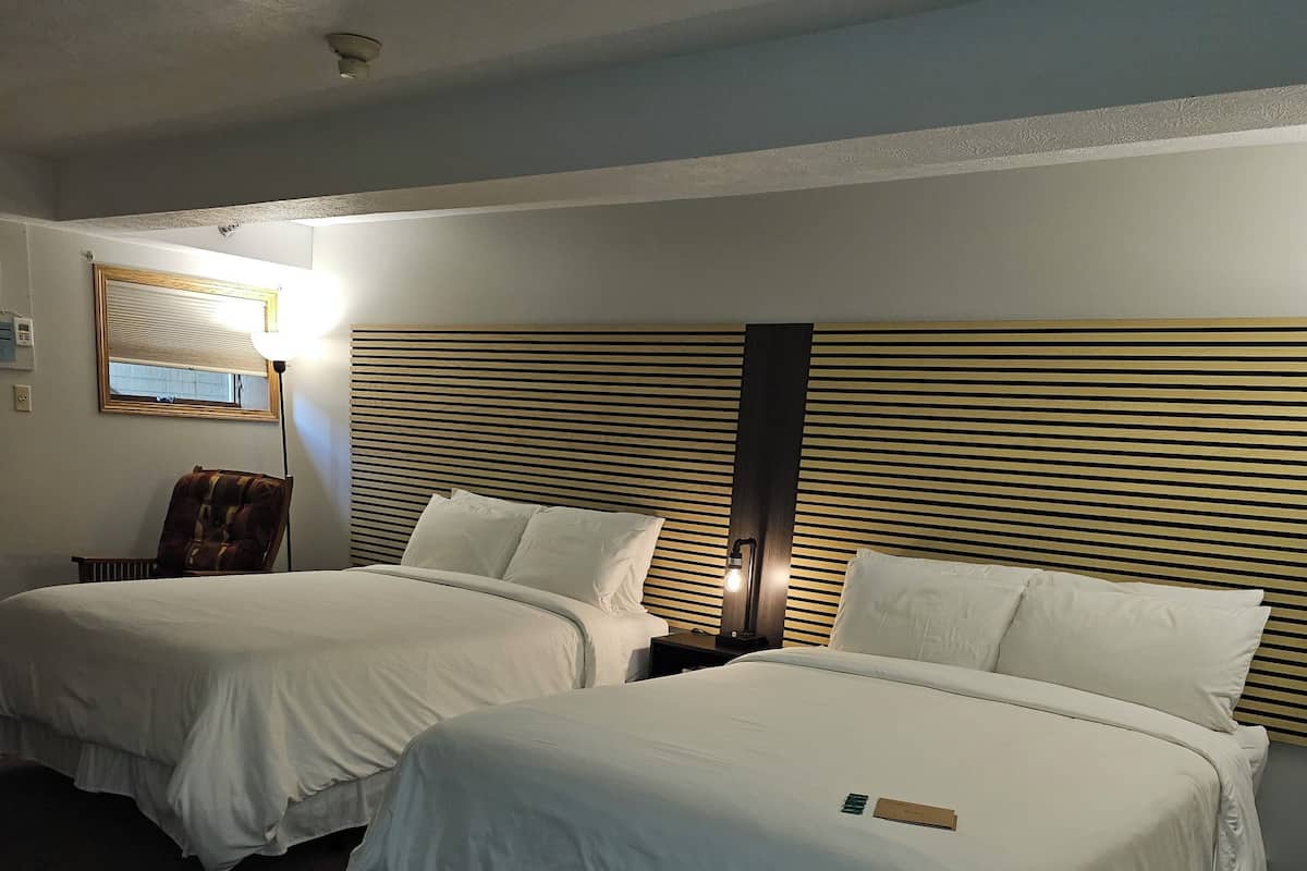 Una habitación de hotel con dos camas, una silla, una mesita de noche, una lámpara y un espejo.