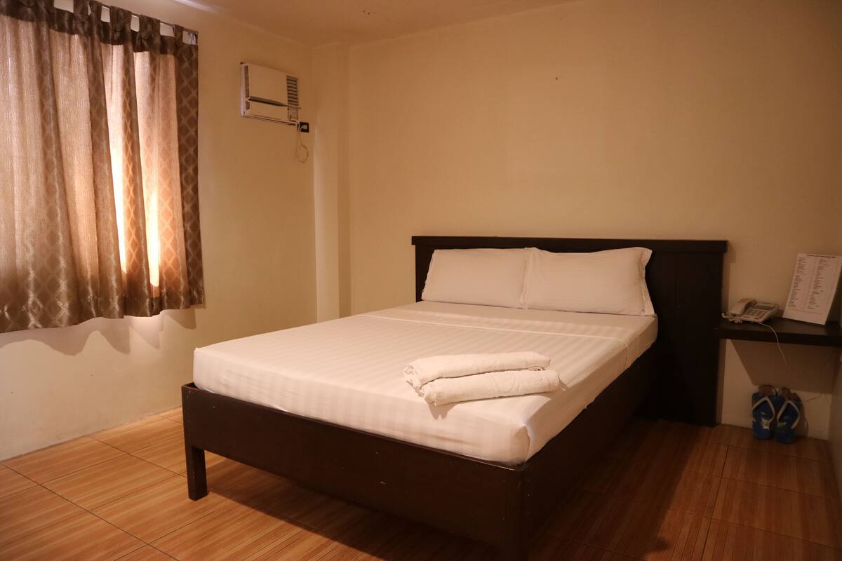 A neatly made bed with white linens, a dark wooden headboard, and a pair of shoes on a small shelf beside the bed.