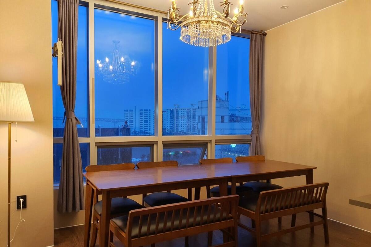 A dining area with a wooden table and benches, large windows with a city view, and a chandelier.