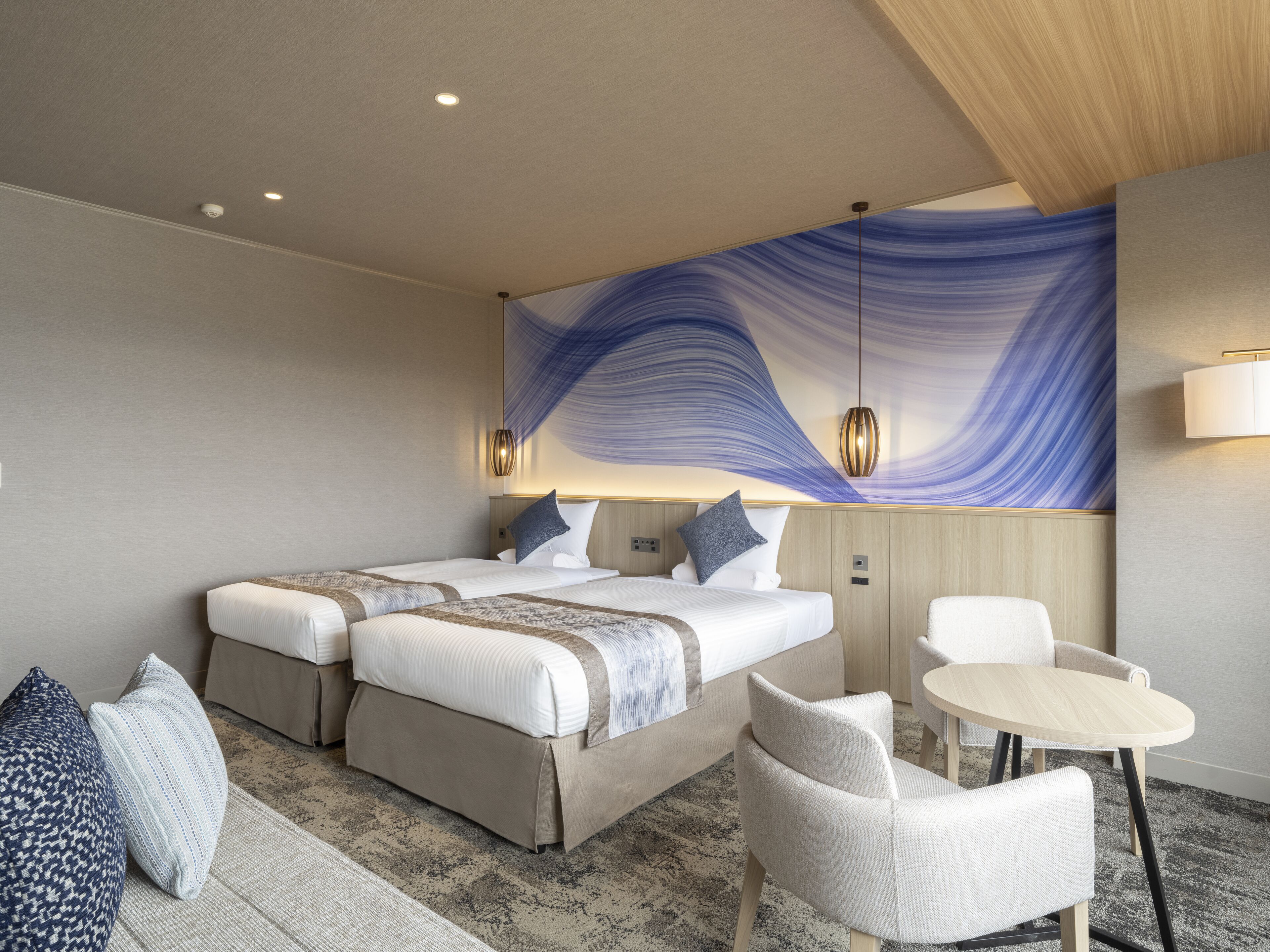 A modern hotel room with two beds, a sitting area with a table and chairs, and a large abstract painting on the wall.