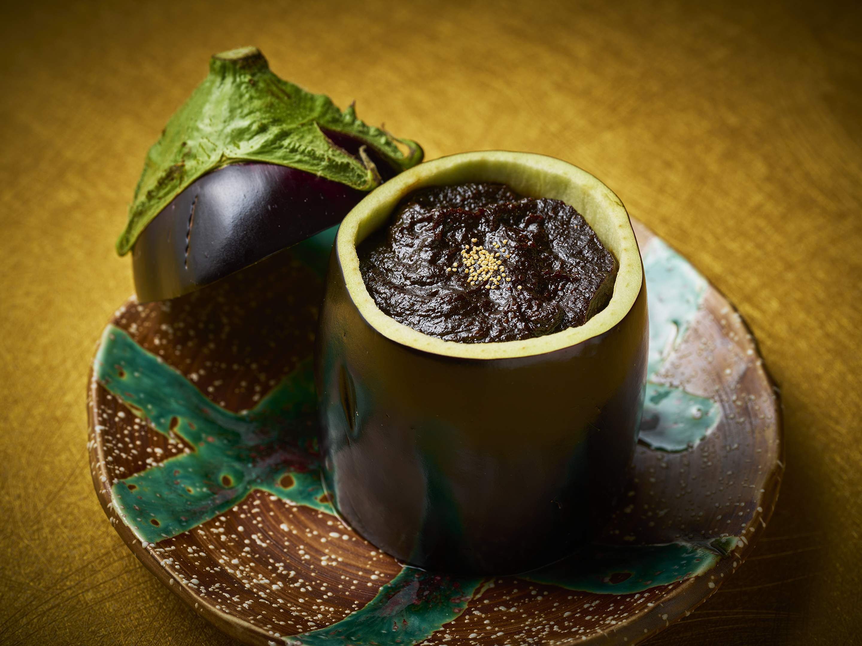 Eine Keramiktasse mit einer dunklen Substanz, vermutlich ein Dessert, auf einem verzierten Teller mit einem Blatt und einer Aubergine.
