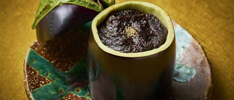 Eine Keramiktasse mit einer dunklen Substanz, vermutlich ein Dessert, auf einem verzierten Teller mit einem Blatt und einer Aubergine.