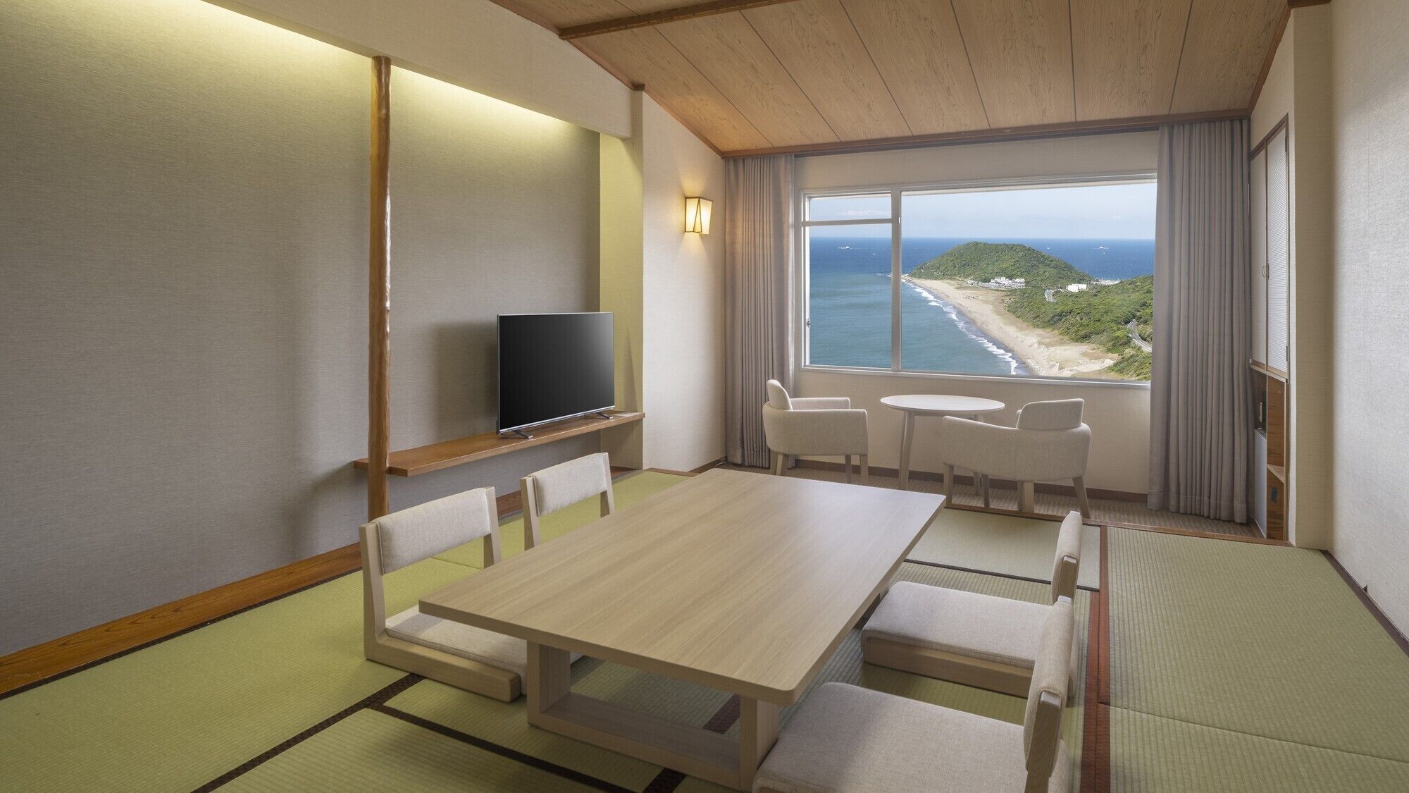 A room with a wooden table, chairs, a flat-screen TV, and a view of the ocean.