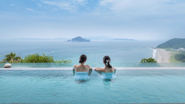 Zwei Personen in einem Infinity-Pool mit Blick auf das Meer und entfernte Inseln.