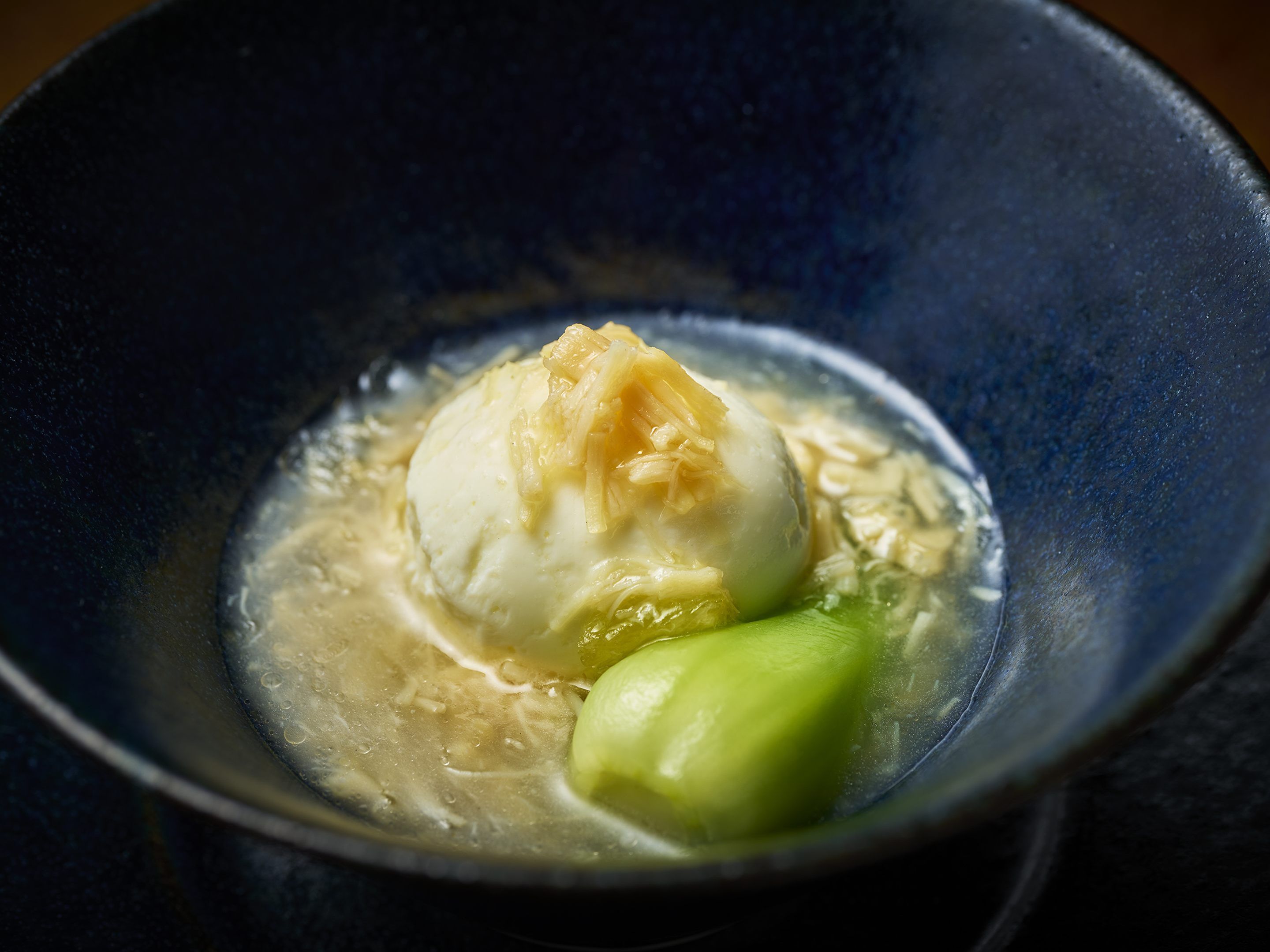 A bowl with a dumpling and a green vegetable, possibly bok choy, in a dark blue bowl.