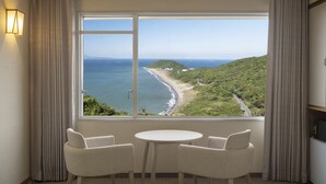 Ein Zimmer mit großem Fenster mit Blick auf Strand und Meer, zwei Stühle und ein kleiner Tisch.