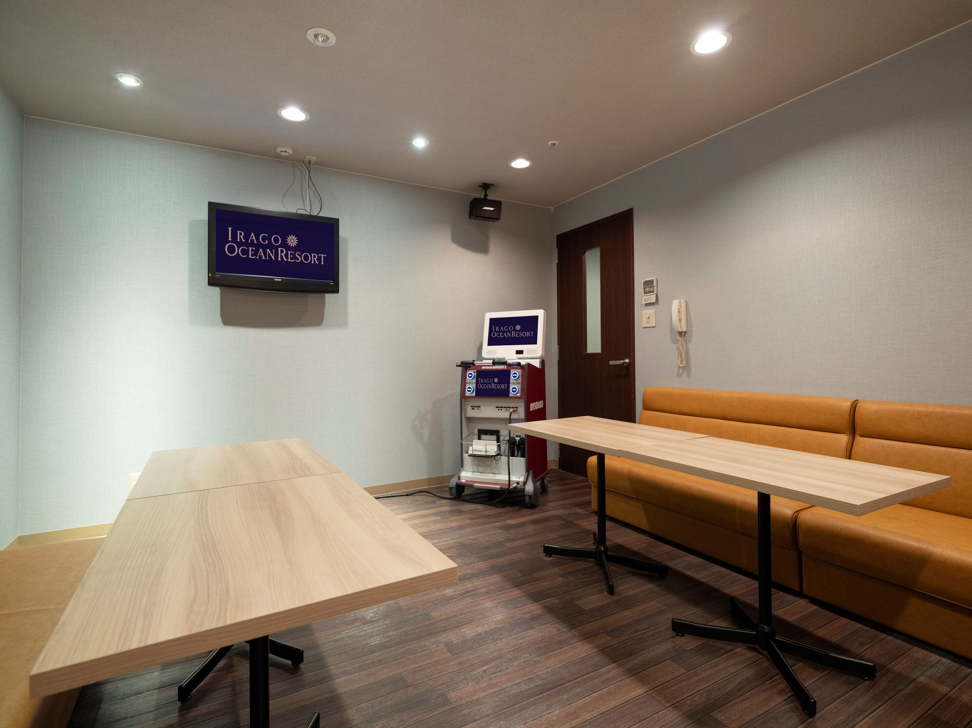 A hotel room with a wooden table, a brown leather couch, a TV screen displaying 'IRAGO OCEAN RESORT', and a minibar.