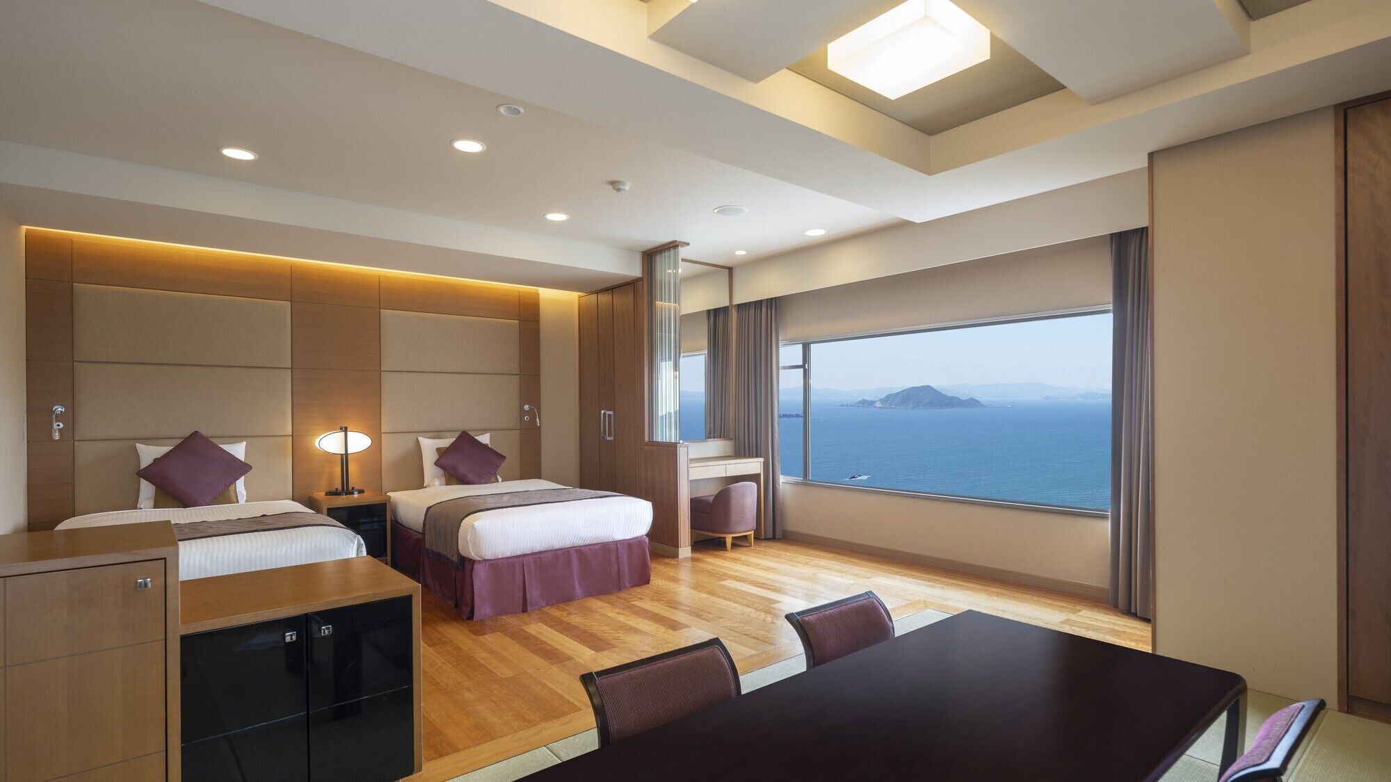A hotel room with two beds, a desk, a chair, a table, and a large window with a view of the sea.