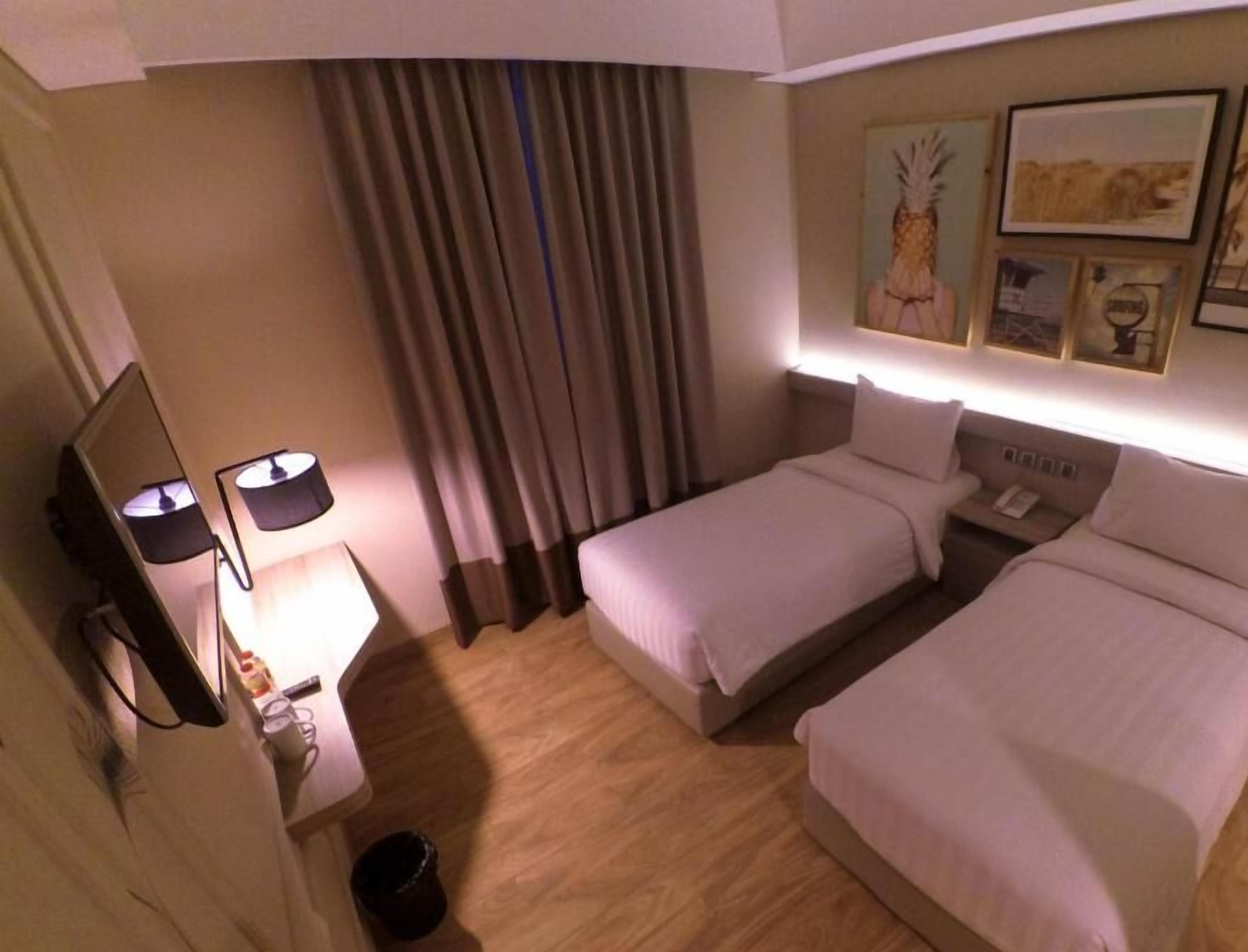 A hotel room with two beds, a desk with a lamp, a trash bin, and a wall with framed pictures.