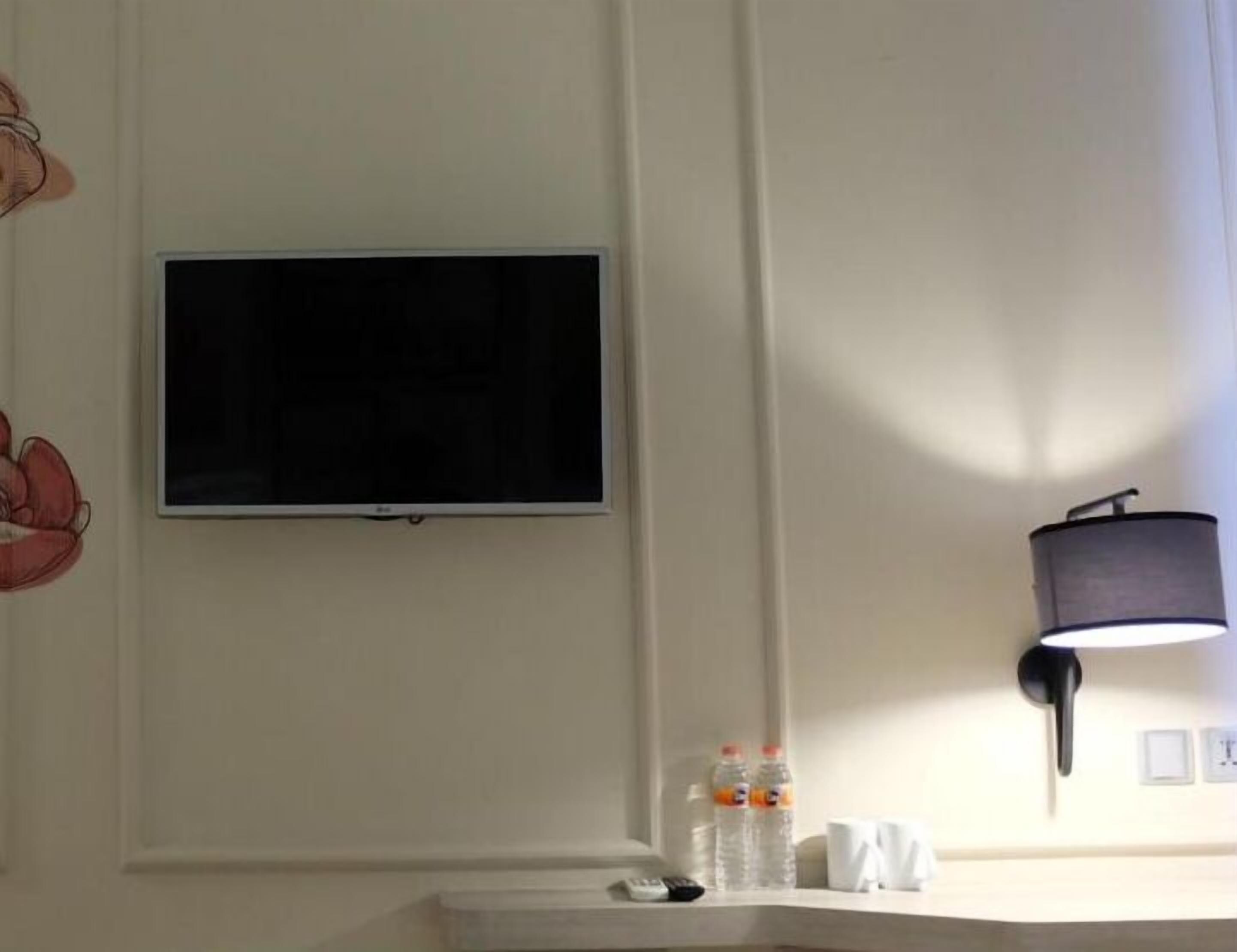 Een hotelkamer met een wandtelevisie, een wandlamp en een aanrecht met waterflessen en een afstandsbediening.