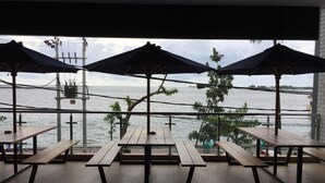 Un espace extérieur aménagé avec des tables en bois et des parasols, offrant une vue sur une étendue d’eau, un pont et des bateaux au loin.