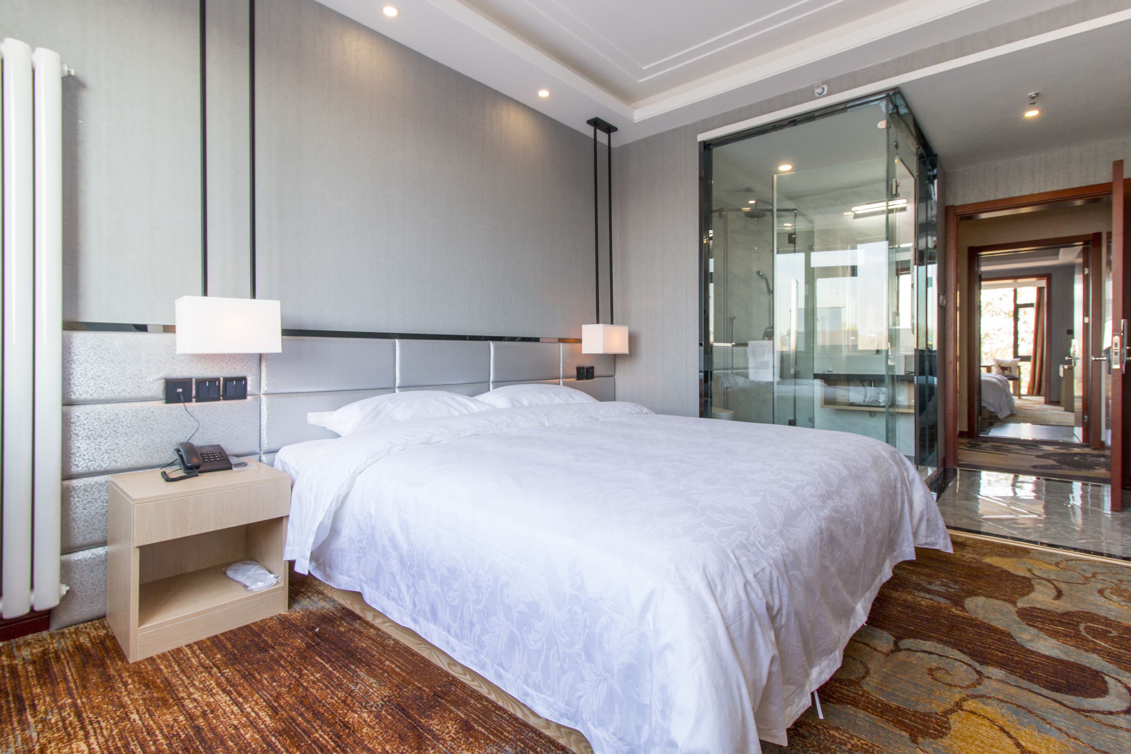 A hotel room with a large bed, bedside tables, a telephone, and a glass wall leading to a bathroom.