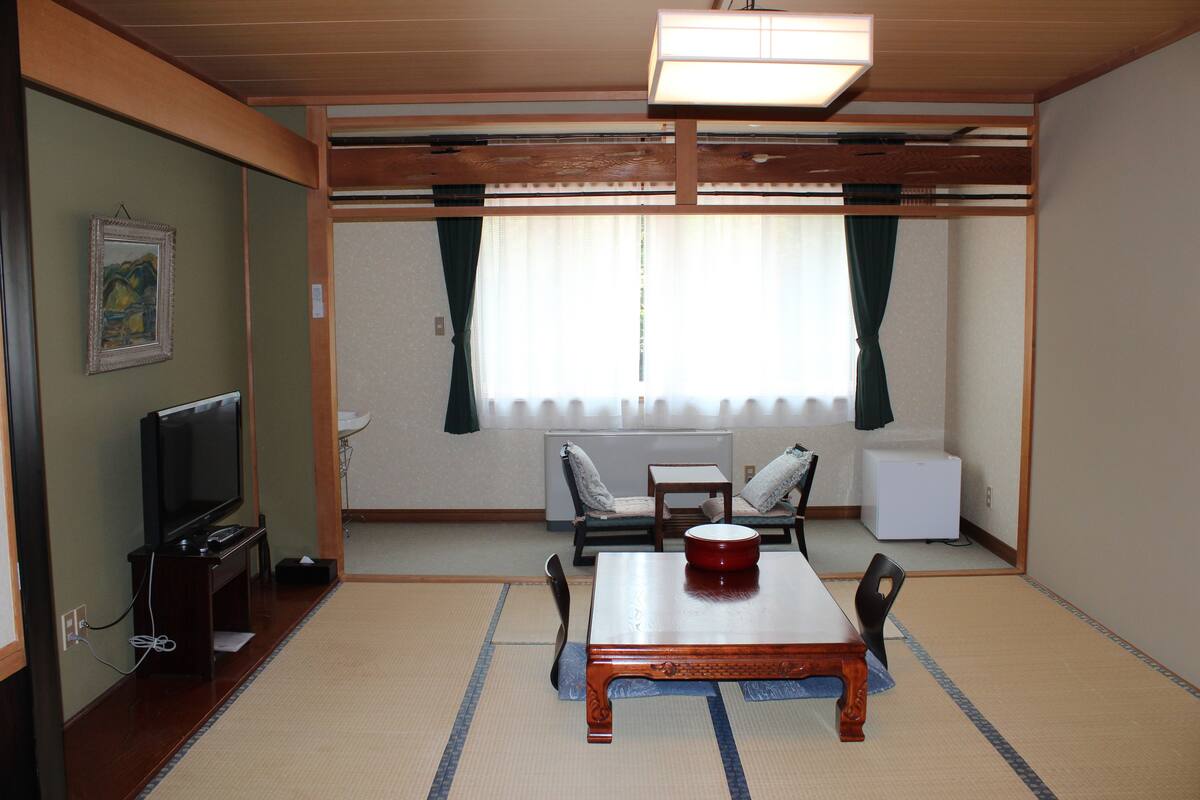Una habitación tradicional japonesa con suelo de tatami, una mesa baja con un cuenco rojo, un televisor y una cama futón.
